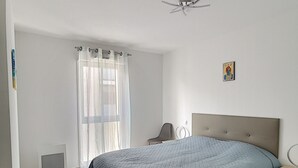 Een slaapkamer met een bed, een stoel, een nachtkastje, een plafondventilator en een raam met gordijnen.