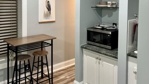 A compact kitchen with a dining area, featuring a wooden table, two stools, a microwave, and a glass shelf with kitchenware. - Thomas Mill Creek Lakefront Property  (Abbeville)