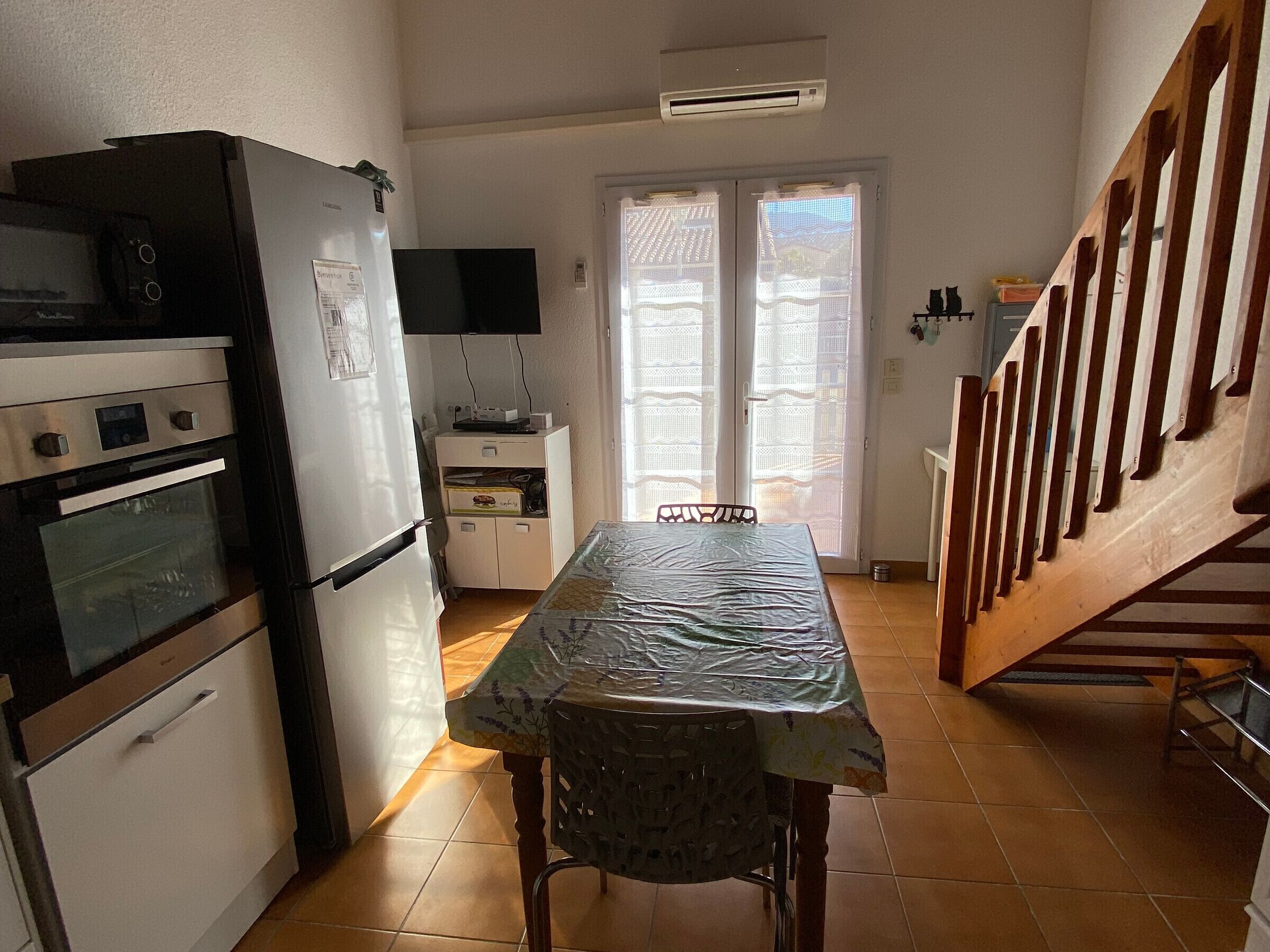 A compact kitchen with a refrigerator, oven, and microwave. There is a dining table with a plastic tablecloth and a chair. A television is mounted on the wall, and a staircase is visible to the right.