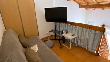 Een kamer met een bank, een televisie op een standaard, een kleine tafel met een bloevas en een raam met gordijnen.