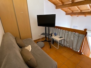 A room with a sofa, a television on a stand, a small table with a vase of flowers, and a window with curtains.
