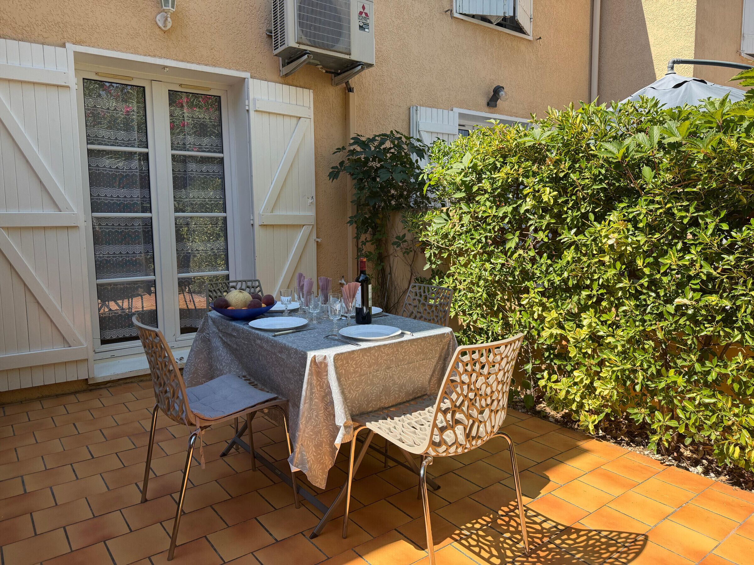 An outdoor dining area with a table set for two, surrounded by a brick patio and a hedge.