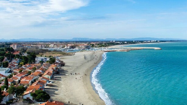 Uma cidade costeira com uma praia, edifícios com telhados de terracota e um céu azul límpido.