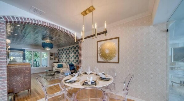 Zona pranzo con tavolo rotondo apparecchiato per quattro persone, sedie trasparenti, un lampadario e carta da parati decorata.