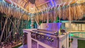 Cocina exterior con parrilla, rodeada de plantas de bambú y techo de paja.