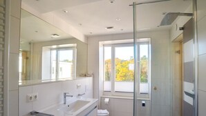 Een moderne badkamer met een glazen douchecabine, een grote spiegel en een raam met uitzicht op herfstkleurige bomen.