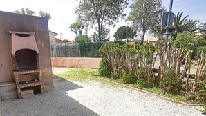 A gravel patio with a brick barbecue area and a small brick oven, surrounded by greenery and a fence.