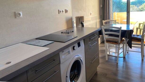 A modern kitchen with a built-in washing machine, a dining area with a table and chairs, and a large window offering a view of the outdoors. - Bright studio with balcony, Wi-Fi, air conditioning, near park (Balaruc-les-Bains)