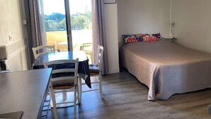 A room with a bed, a table with chairs, a window with curtains, and an air conditioning unit. - Bright studio with balcony, Wi-Fi, air conditioning, near park (Balaruc-les-Bains)