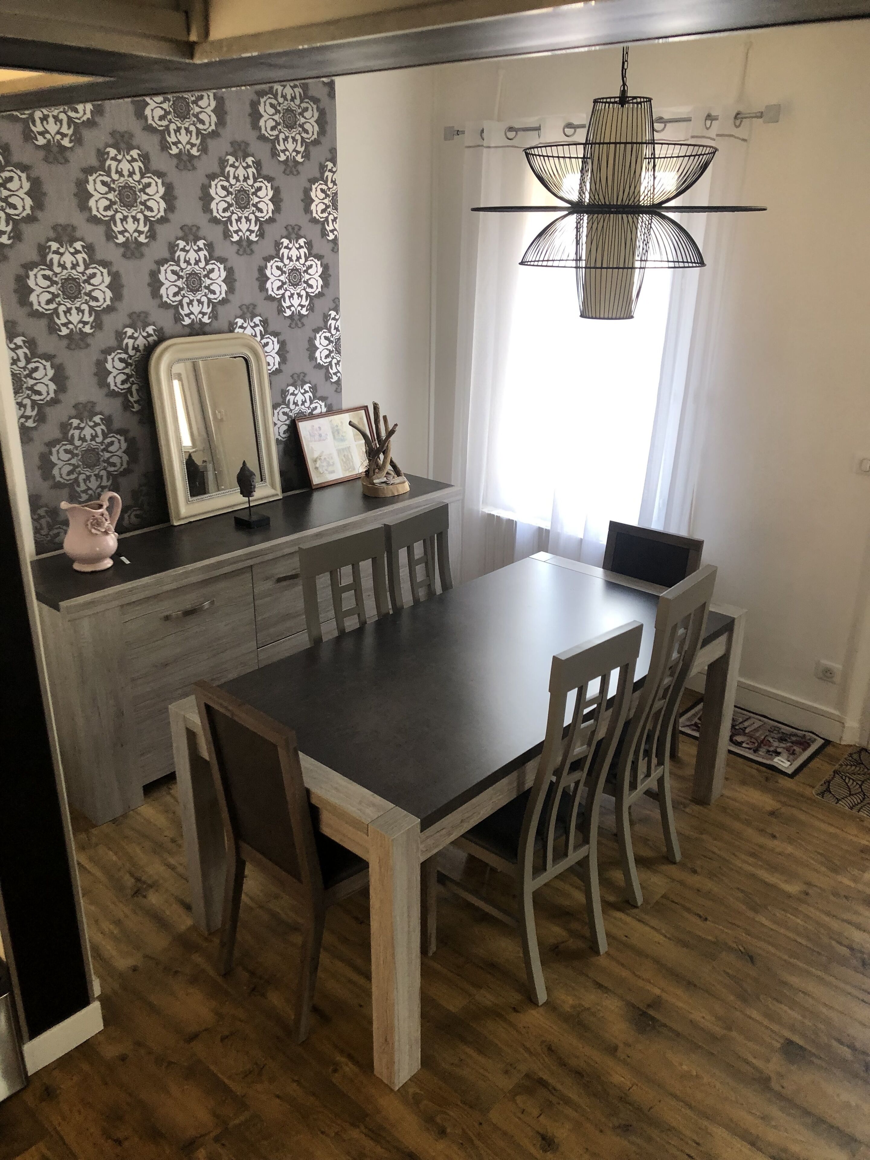 A dining area with a wooden table and chairs, a chandelier, a mirror, and a patterned wallpaper.