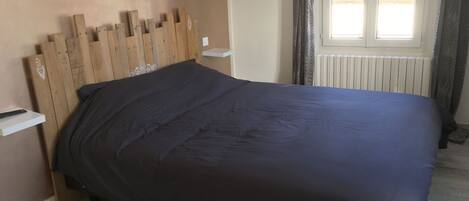 A bedroom with a wooden headboard, a dark bedspread, a white bedside table, and a window with curtains.