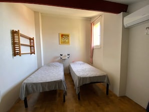 A small room with two single beds, a wooden ladder, a painting on the wall, and a window with curtains.
