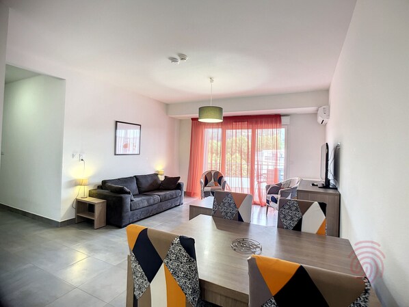 Una sala de estar moderna con un sofá, una mesa de comedor y sillas. Hay una lámpara, un cuadro en la pared y una ventana con cortinas.