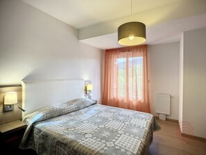 Un dormitorio con cama, lámparas de noche, una ventana con cortinas y un radiador.