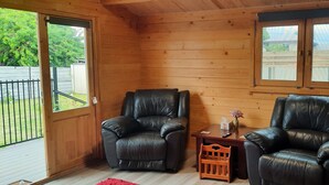 A wooden cabin interior with two leather chairs, a small table, and a window with a view of a fence and greenery.