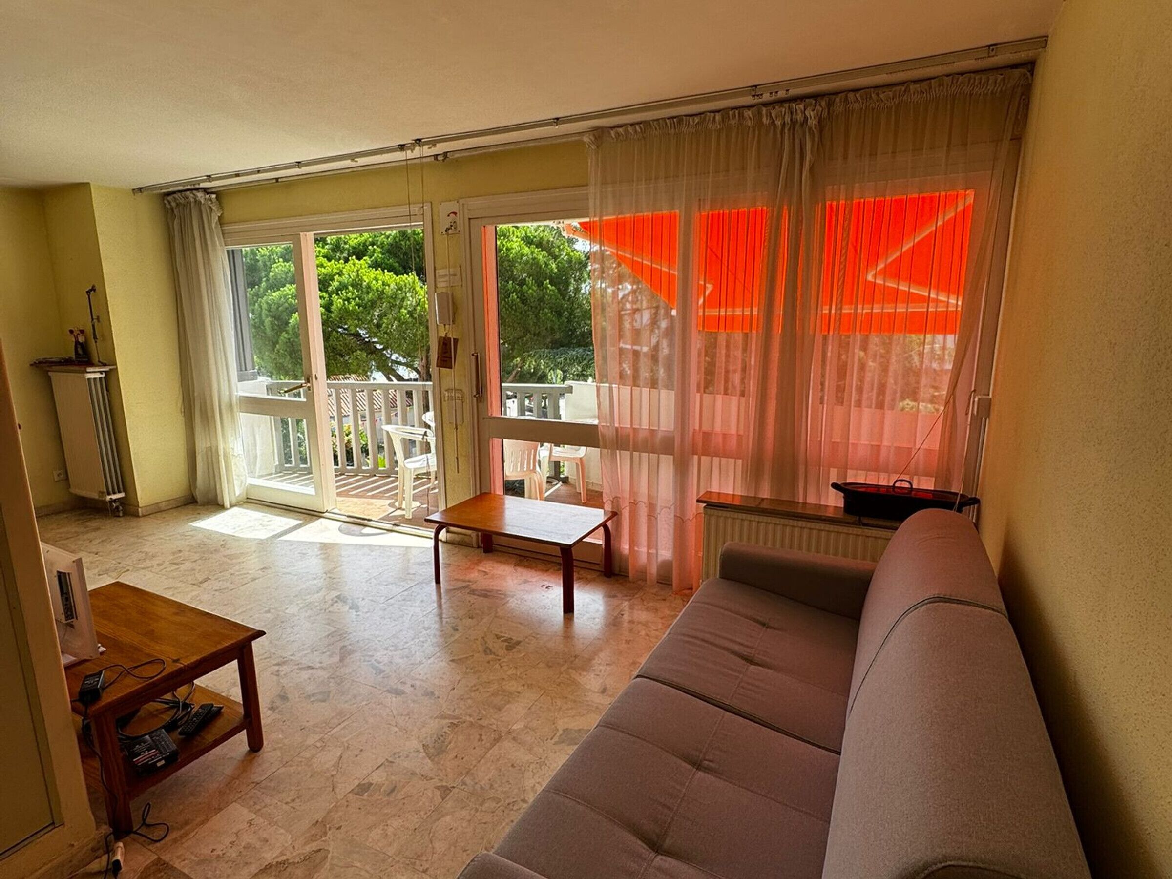 A living room with a sofa, a table, and a radiator. There is a balcony with a view of trees and a railing.