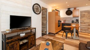 Un espace de vie compact comprenant un téléviseur posé sur un meuble en bois, un coin cuisine équipé d’appareils modernes et une table à manger avec des chaises.