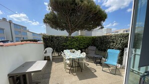 Une terrasse avec une table et des chaises, une chaise bleue, un arbre et une haie.