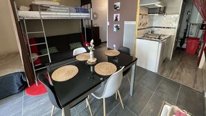 Un espace de vie compact comprenant un lit superposé, une petite kitchenette et un coin repas avec une table noire et des sets de table en fibres tressées.