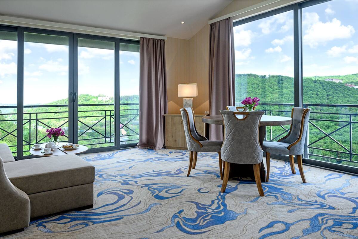 A hotel room with a balcony, a sofa, a table with chairs, and a view of a green landscape.
