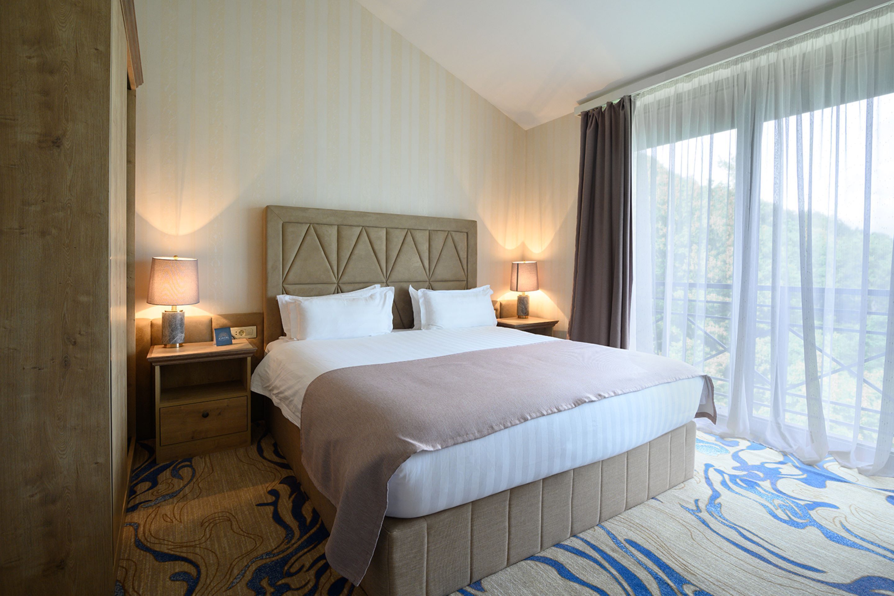 A hotel room with a large bed, two bedside tables with lamps, a window with curtains, and a carpet with a blue pattern.