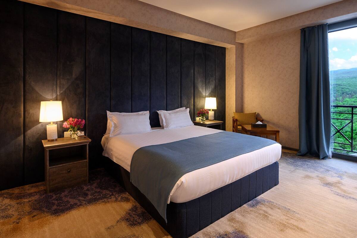 A large bed with a dark headboard, two bedside tables with lamps, and a view of greenery through a window.