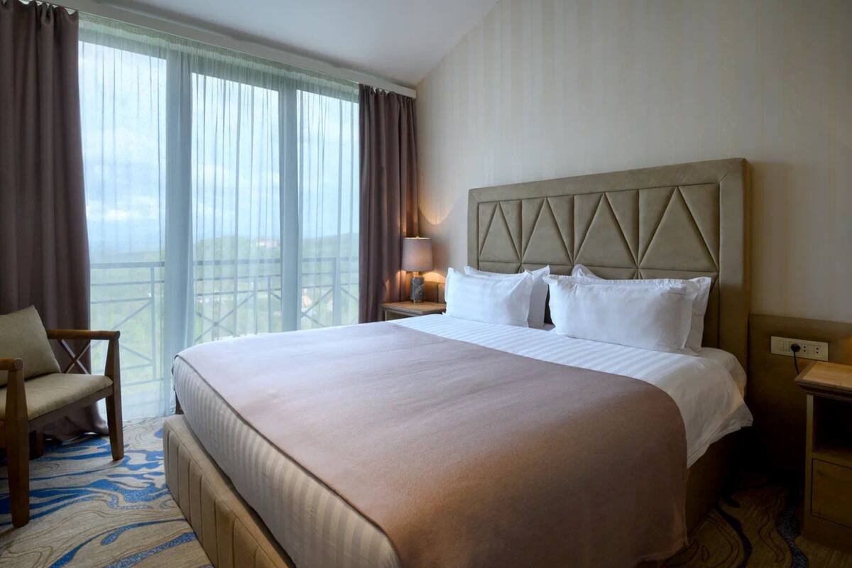 A hotel room with a large bed, a chair, a nightstand with a lamp, and a view through the window.