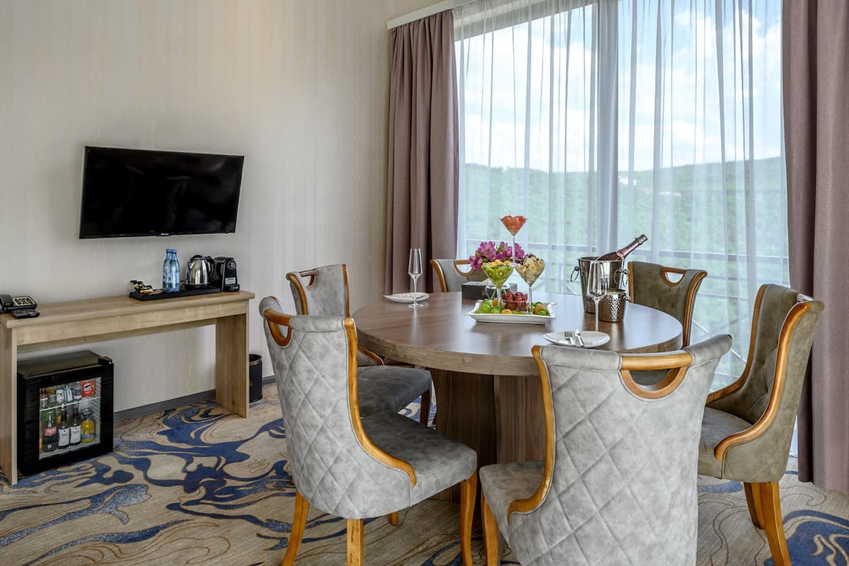 A hotel dining area with a round table, four upholstered chairs, a television on the wall, and a view of the outdoors through large windows.