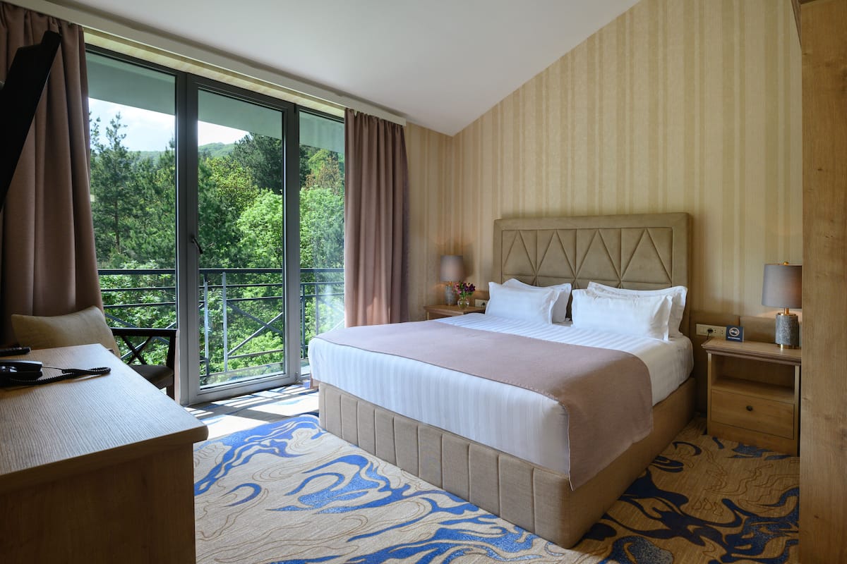 A hotel room with a large bed, a desk, and a view of greenery through the window.