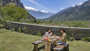 A picnic table with a stone wall backdrop, surrounded by lush greenery and mountains in the distance. - Welcomhotel By ITC Hotels Hamsa Manali (Manali)