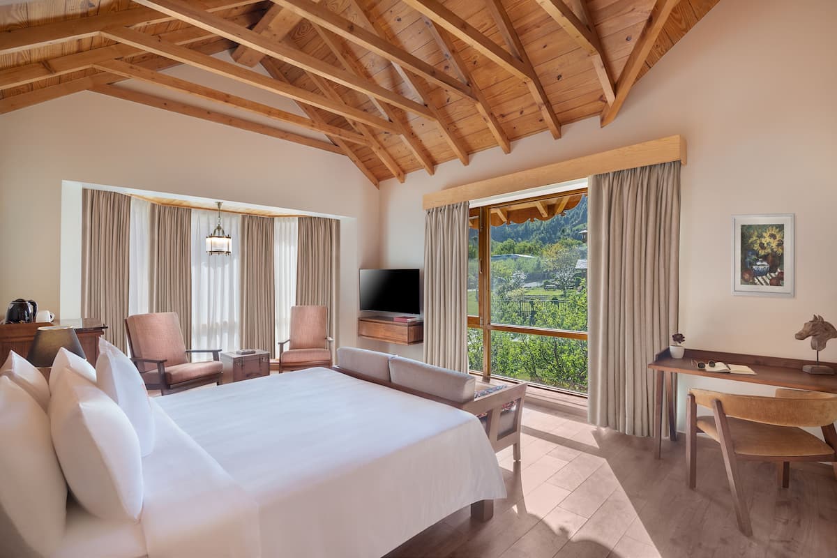 Ein Hotelzimmer mit einem großen Bett, einem Schreibtisch, einem Sessel und einem Fernseher, mit Blick ins Freie.