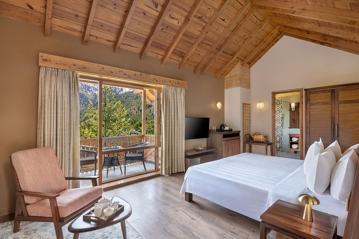 Ein Schlafzimmer mit einem großen Bett, einem Fernseher und einem Schreibtisch. Ein Sessel und ein Tisch mit Tablett sind ebenfalls vorhanden. Das Zimmer verfügt über Holzbalkendecken und bietet durch das Fenster einen Blick auf Bäume und Berge.