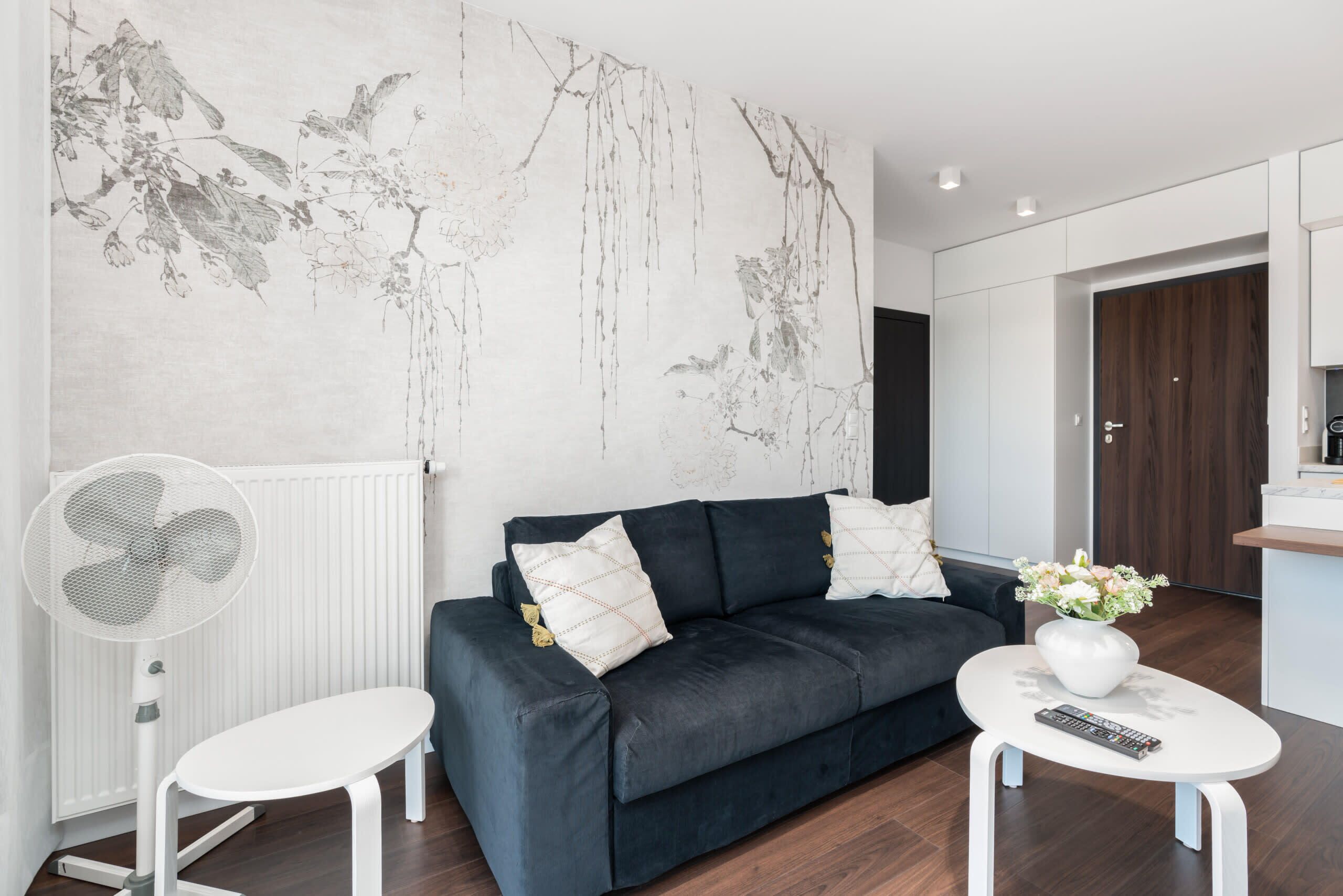 Un salon moderne avec un canapé gris foncé, des coussins blancs, un ventilateur blanc, une petite table blanche ornée d’un vase de fleurs et un mur sur lequel est accrochée une grande peinture abstraite.