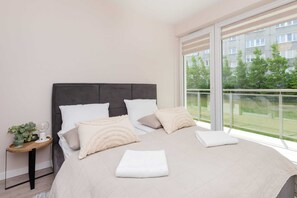 Un dormitorio moderno con una cama grande, una mesita de noche con una planta y vistas a un edificio y árboles por la ventana.