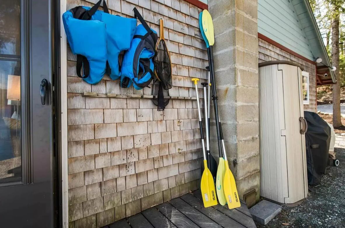 Pared exterior de madera con una puerta, una bolsa azul, una pala y un kayak apoyado en la pared.