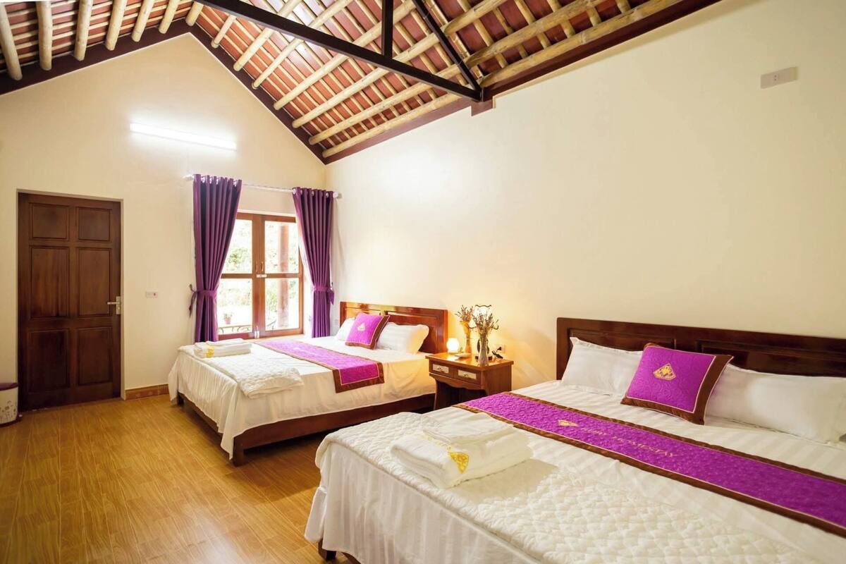 Habitación de hotel con dos camas, piso de madera, techo inclinado y una ventana con cortinas moradas.