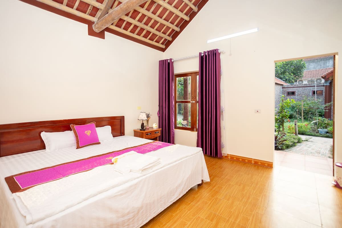 Un dormitorio con techo de madera, cama con ropa de cama blanca y un cojín rosa decorativo, mesita de noche con lámpara, y ventana con cortinas moradas.