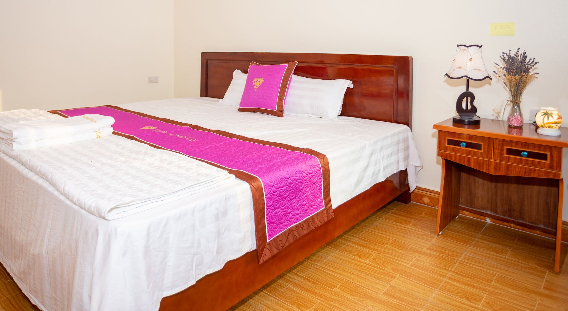 A double bed with white linens and a pink decorative runner, a wooden nightstand with a lamp and a vase of flowers, and a wooden headboard.