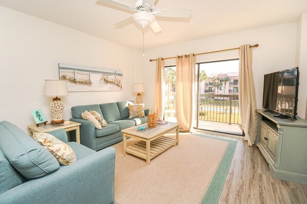 A living room with a sofa, coffee table, and TV stand. There is a ceiling fan, a window with curtains, and a view of buildings outside. - Ocean Village Club Q22 (St. Augustine)