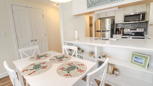 A compact kitchen with white cabinetry, a stainless steel refrigerator, a microwave, and an oven. There is a dining table with a floral placemat and white chairs. - Enjoy the Sunrise and the Sunset from your very own Balcony! (St. Augustine)