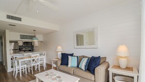 A cozy living room with a beige sofa, white coffee table, and matching side tables. A kitchen with white appliances and a dining area with white chairs is visible in the background. - Enjoy the Sunrise and the Sunset from your very own Balcony! (St. Augustine)