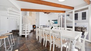 A spacious dining area with a large white table and chairs, a fireplace, and a stone wall. - Ubuntu by Experience Jervis Bay (Vincentia)