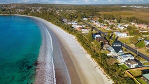 An aerial view of a coastal residential area with houses along the beach, a clear sky, and a sign for Jervis Bay. - Ubuntu by Experience Jervis Bay (Vincentia)