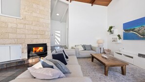 A modern living room with a stone fireplace, a sofa with striped cushions, a wooden coffee table, and a painting of a coastal landscape. - Ubuntu by Experience Jervis Bay (Vincentia)