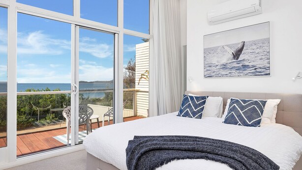 A bedroom with a large bed, a view of the sea, and a balcony with chairs. - Ubuntu by Experience Jervis Bay (Vincentia)