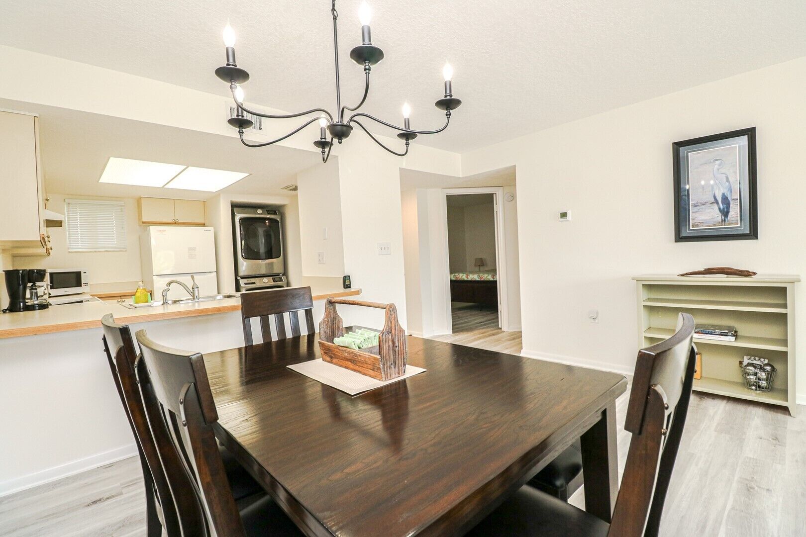 A dining area with a dark wooden table, six chairs, a chandelier, a kitchen with a microwave, oven, and sink, and a framed picture on the wall.