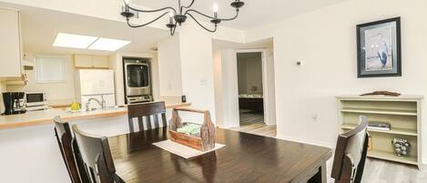 A dining area with a dark wooden table, six chairs, a chandelier, a kitchen with a microwave, oven, and sink, and a framed picture on the wall.