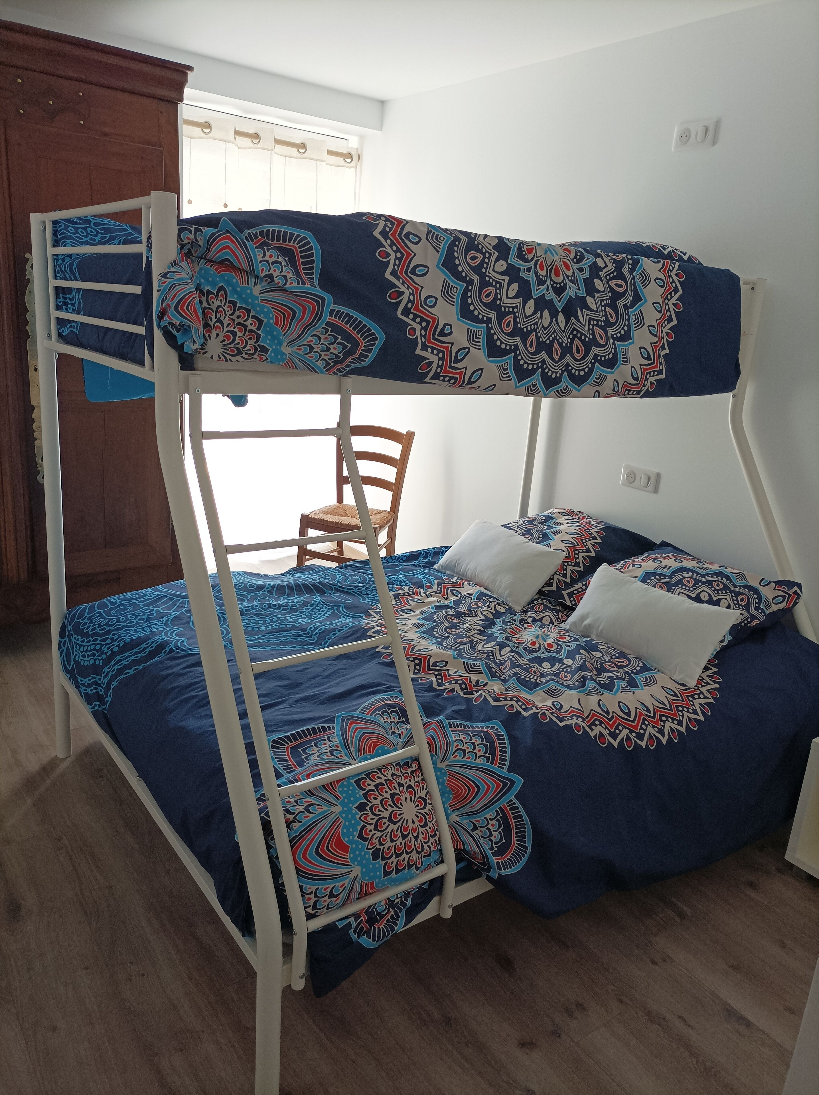 A bunk bed with a patterned bedspread and pillows, a wooden chair, and a window with curtains.