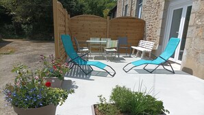 Une terrasse extérieure avec deux chaises longues, une table et des chaises. On aperçoit, à l’arrière-plan, des plantes en pot et une clôture en bois.