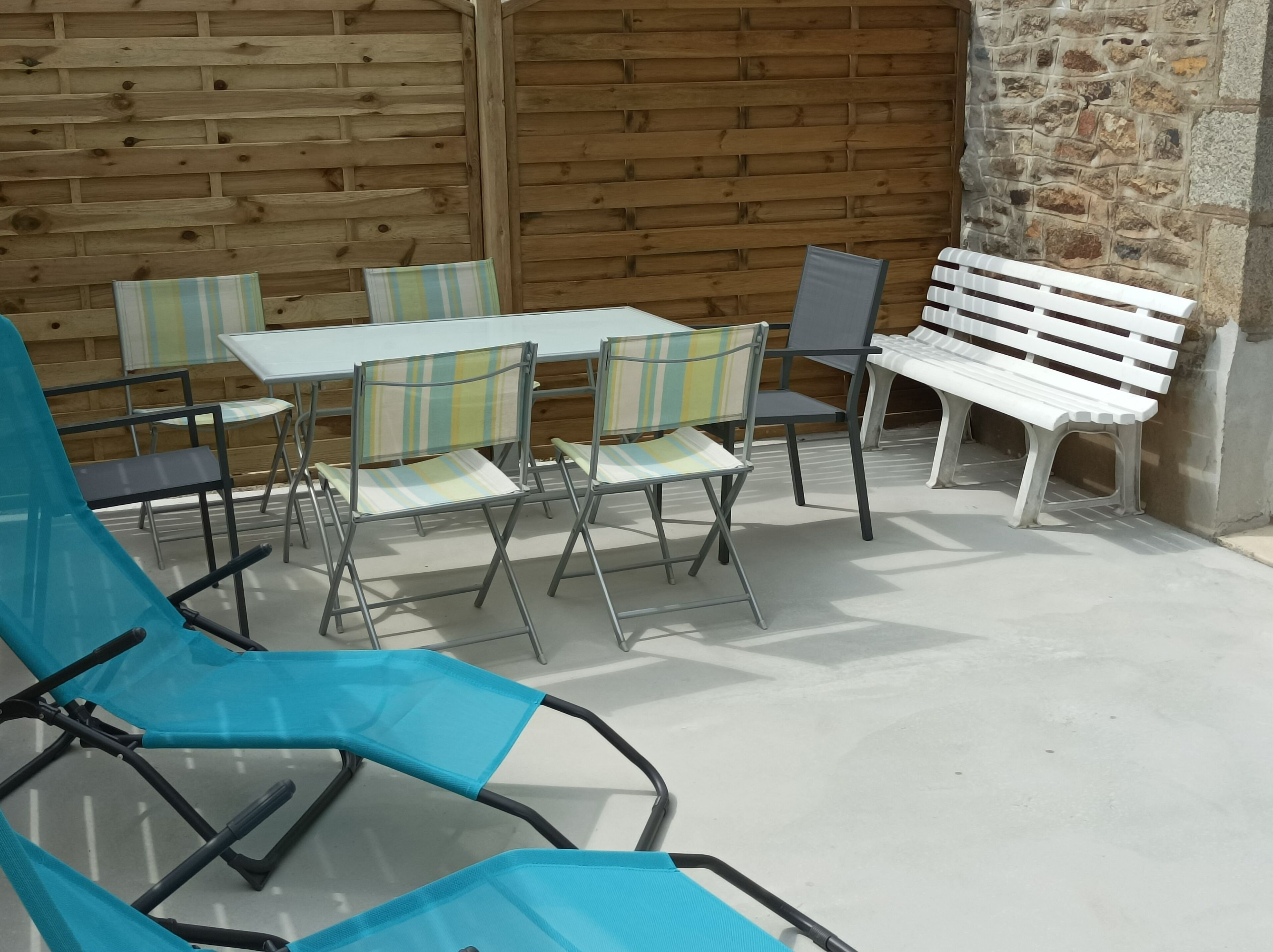 Un espace extérieur aménagé avec des chaises en métal et un banc blanc, adossé à un mur en bois et à un mur en pierre.